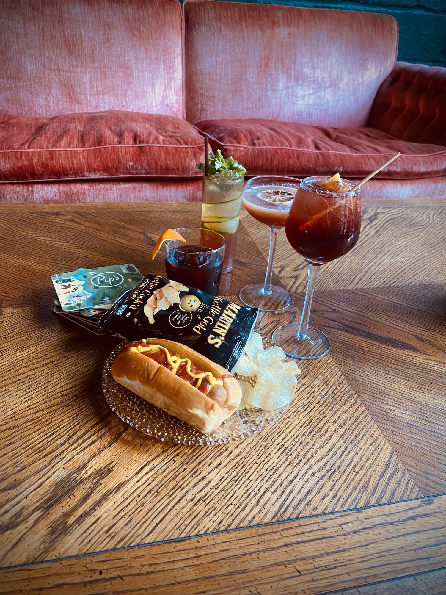 A plate with a hot dog and potato chips sits on a wood coffee table, surrounded by craft cocktails, with a comfortable couch in the background. 