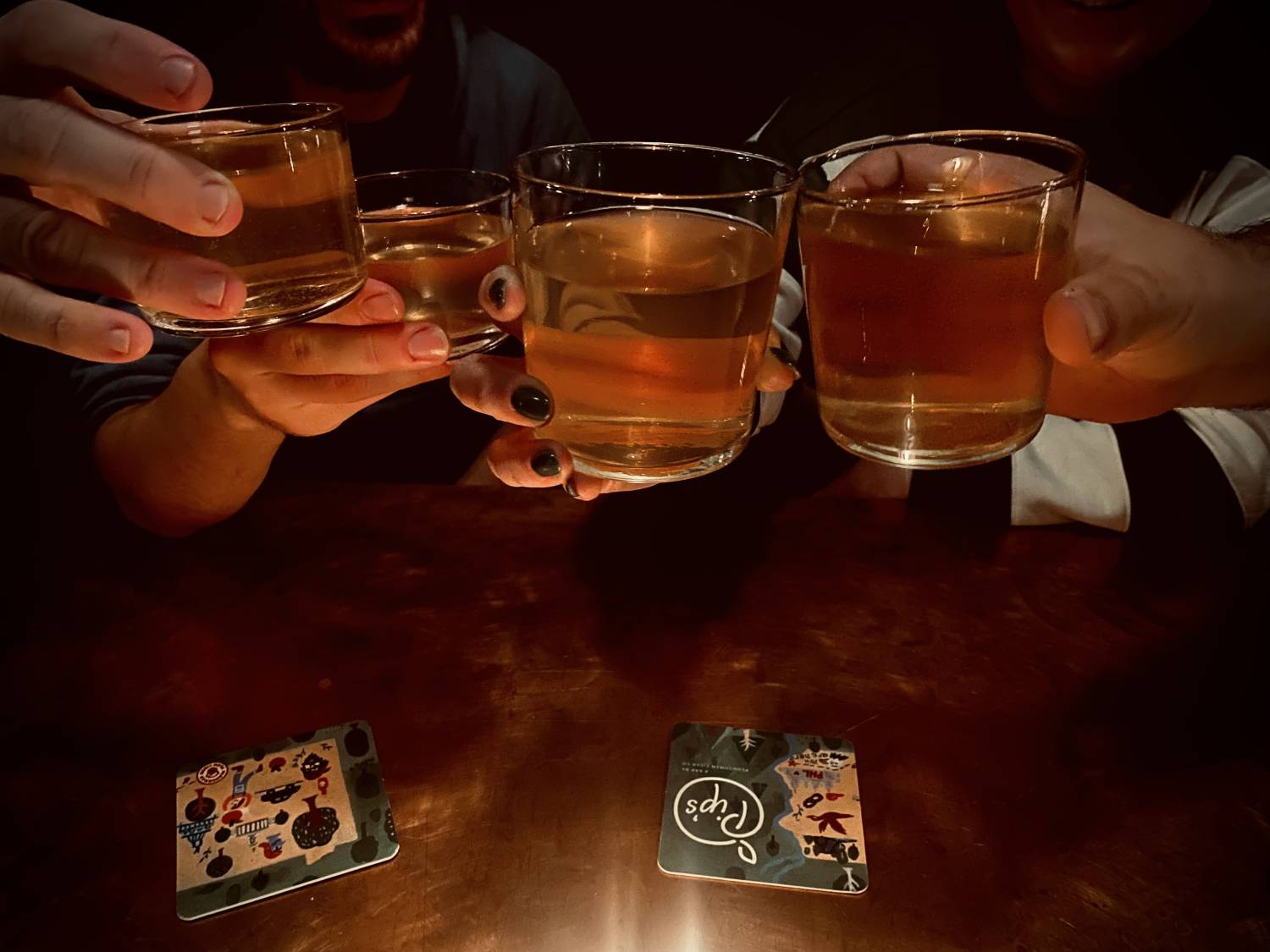 Close-up of hands holding glasses of craft cider in a "cheers" gesture in a dark, cozy bar.